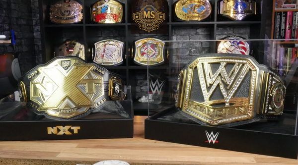 A championship belt display featuring the NXT Championship and WWE Championship belts, with several other championship belts on shelves in the background.